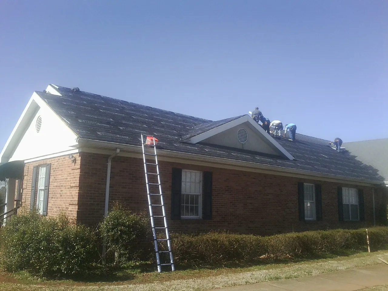 Crew tearing shingles off a brick ranch roof with a ladder set — Emergency Roof Repair work by our crew in Wilmot