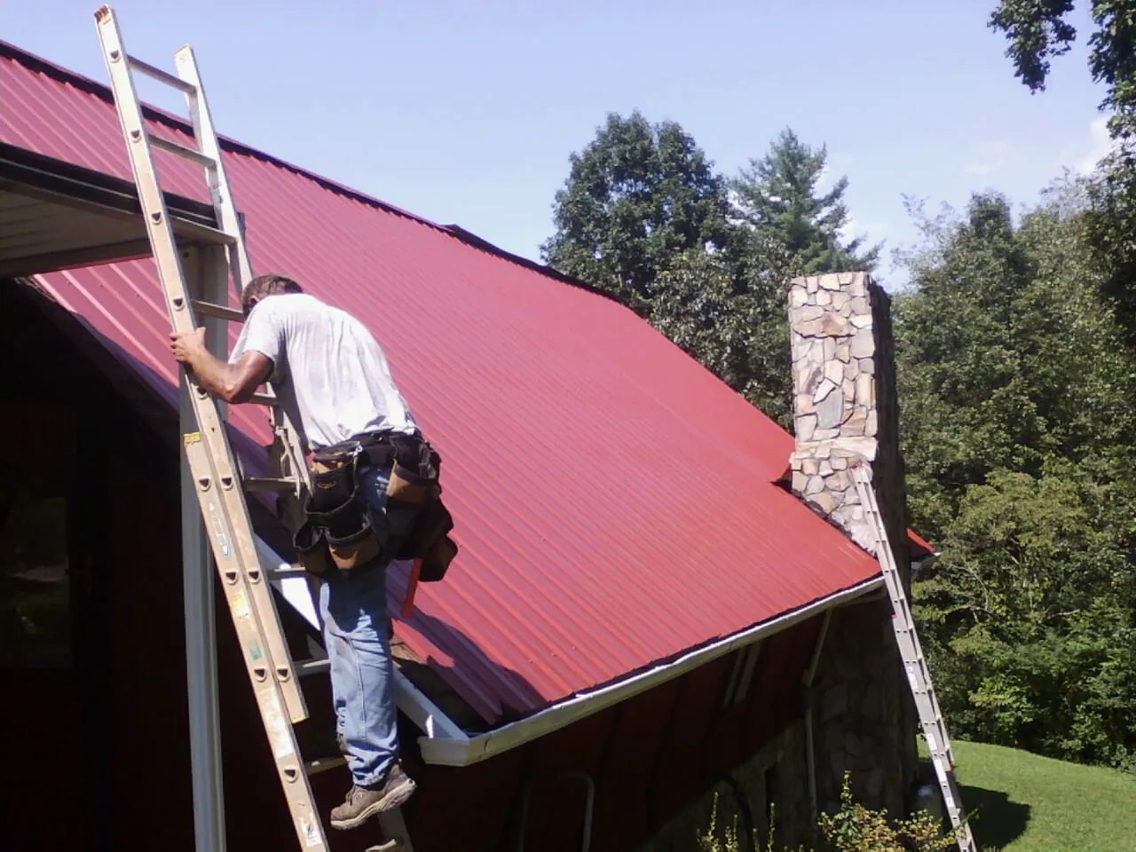Roofer on a ladder beside a red metal roof and stone chimney — Soffit & Fascia Repair by Roofing Brothers in Wilmot